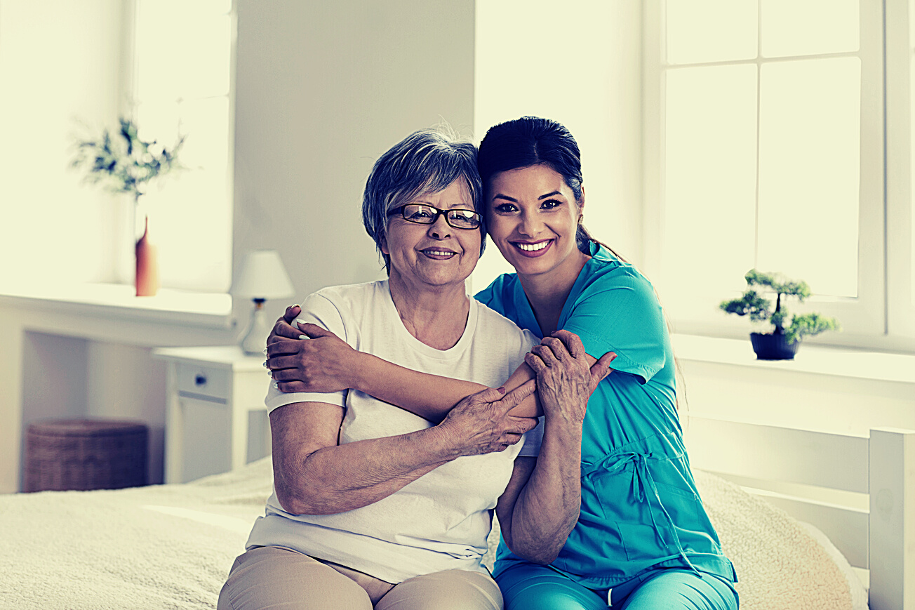 Portrait of Happy Caregiver Together with Senior Patient Sitting on Bed in Retirement Home